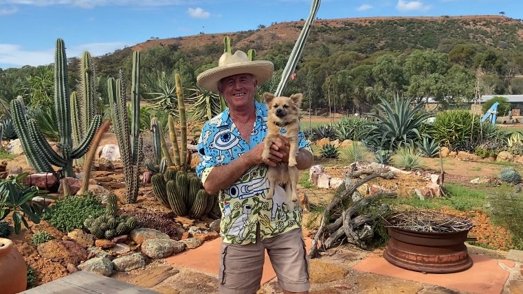 A man holding a small dog and wearing a hat stands in front of tall, spiked cacti.