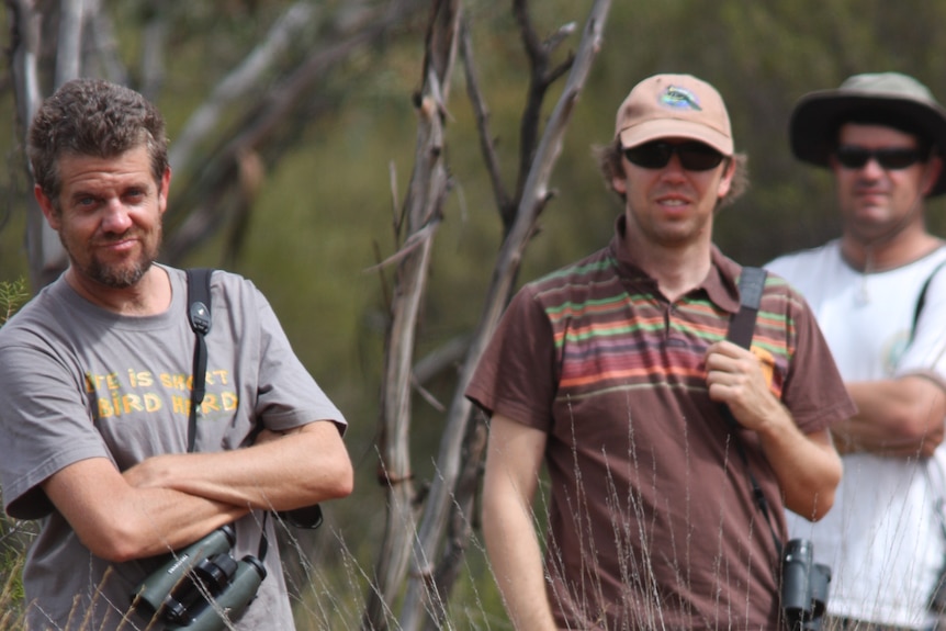 Three men stand in grassland with binoculars handing around their necks, wearing t-shirts.