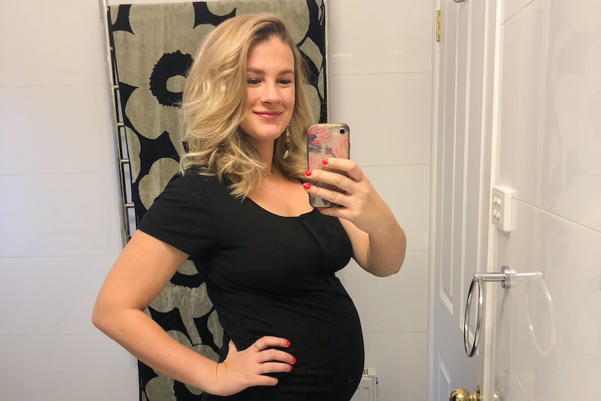 Grace, with blonde hair out, wears black tight dress as she takes a selfie of her pregnancy bump in mirror