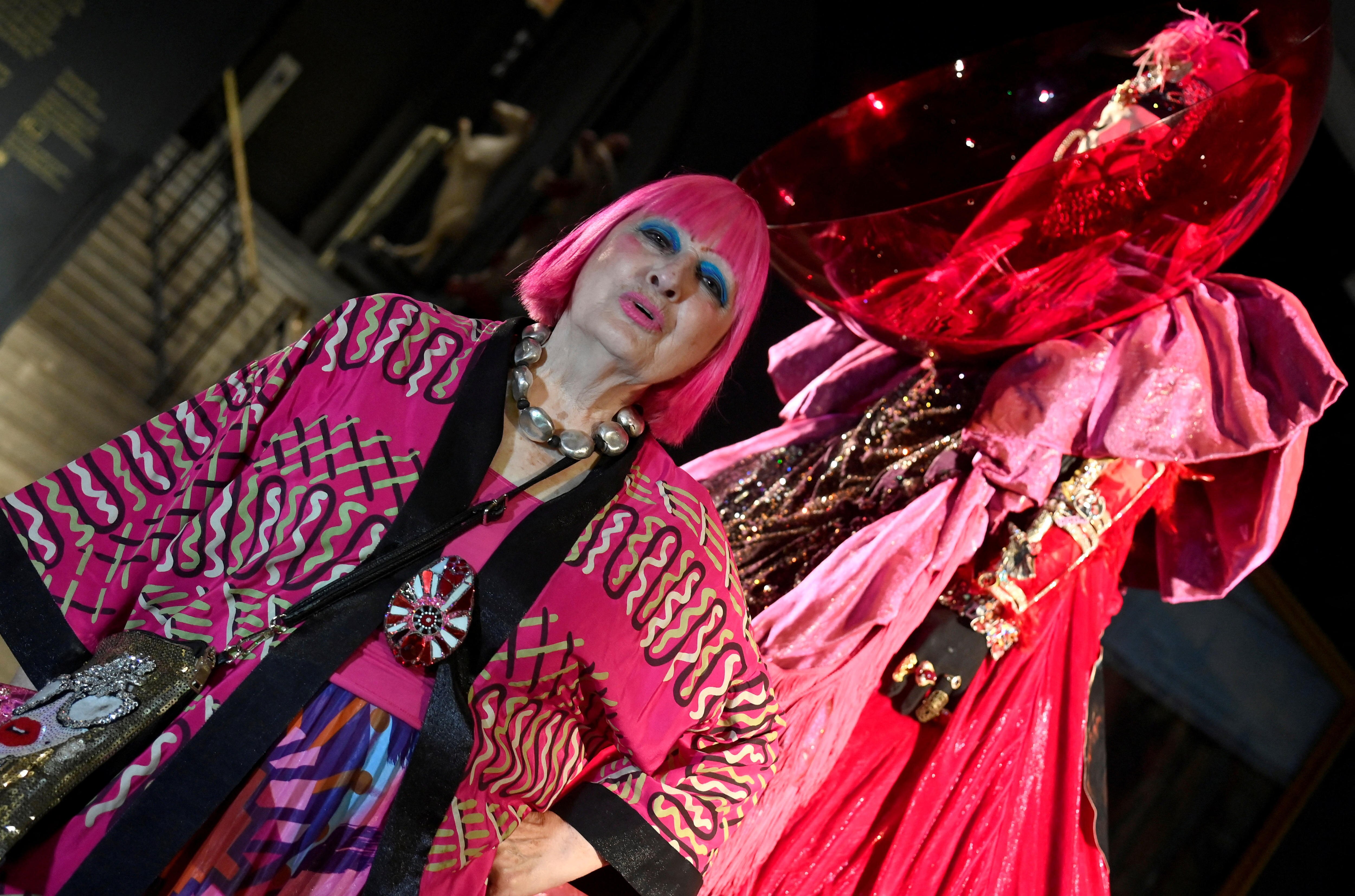 An elderly woman with bright pink hair stands in front of an extravagant red and pink outfit on a mannequin.