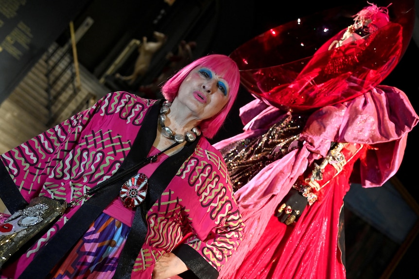 An elderly woman with bright pink hair stands in front of an extravagant red and pink outfit on a mannequin.