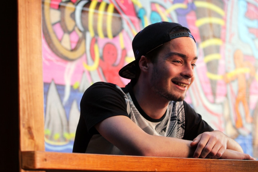 A young man with a cap on backwards looks off to the right of the camera, leaning on a wooden rail.