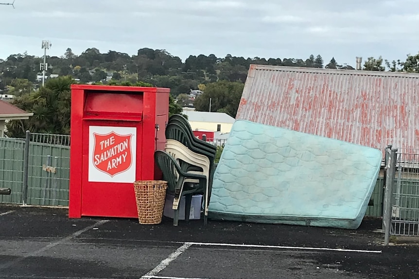 A red donations bin with plastic chairs and a used mattress dumped around it