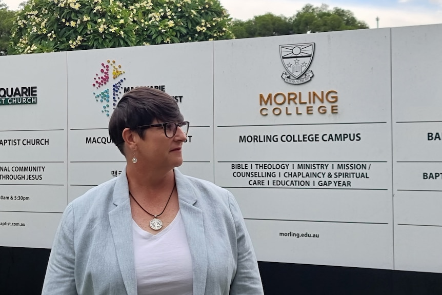 Woman wearing light blue jacket and white shirt standing outside Morling College campus sign.