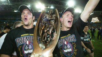 Two NRL players wearing celebratory t-shirts hold the trophy