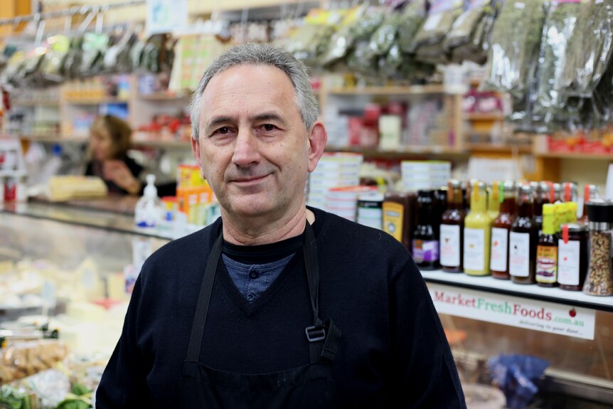 Jim Katsaros smiles, dressed in a jumper and black apron, as he stands in front of a deli counter brimming with Greek foods.