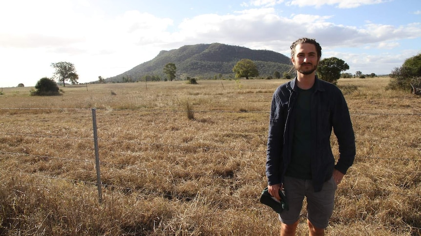 A man stands in an empty paddock with a mountain in the background.