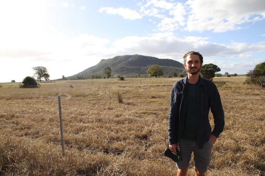 A man stands in an empty paddock with a mountain in the background.