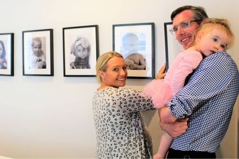 A man holding a toddler stands next to a smiling woman against a wall of photographs of children.
