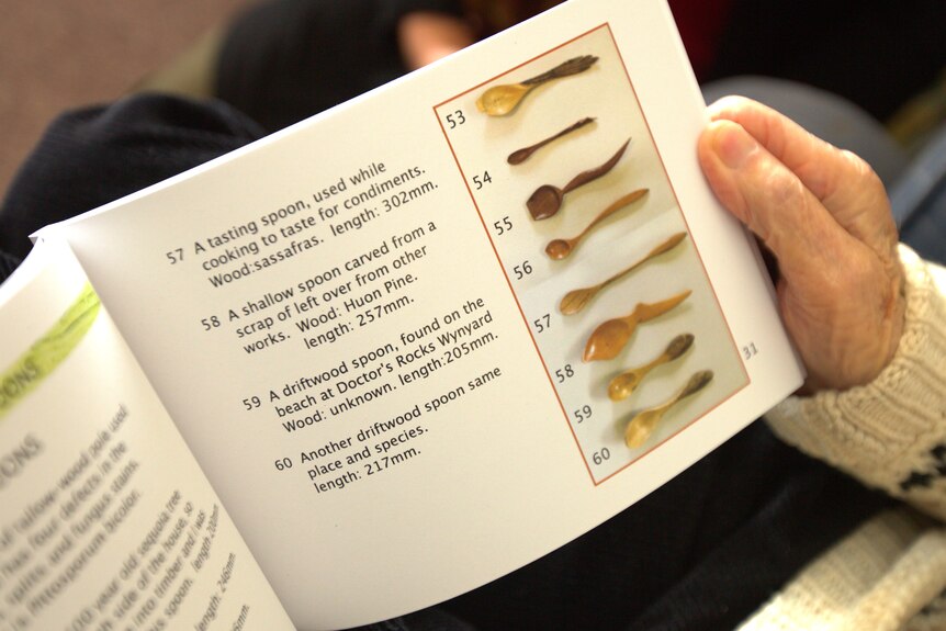 An open book, showing photographs of wooden spoons alongside descriptions of them, held by an old hand.