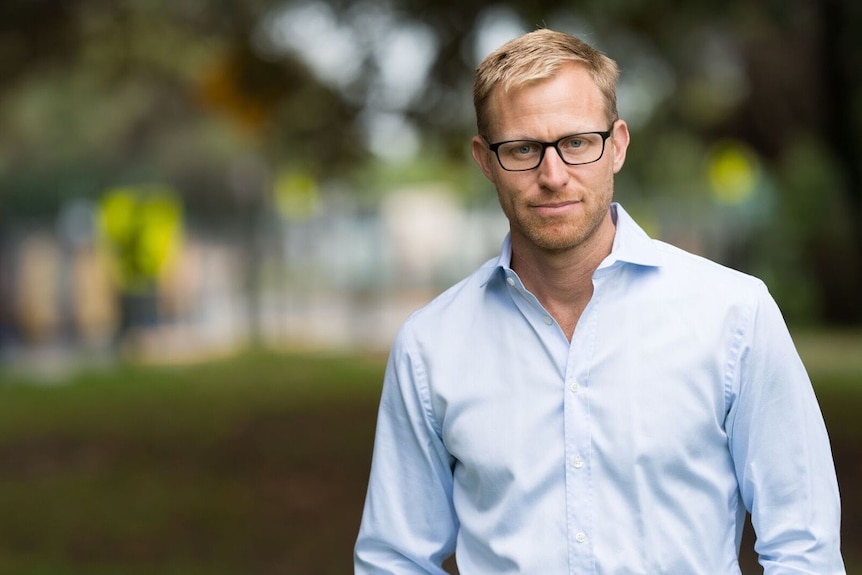 A fair-haired, bespectacled man in a light-coloured business shirt standing in a park.