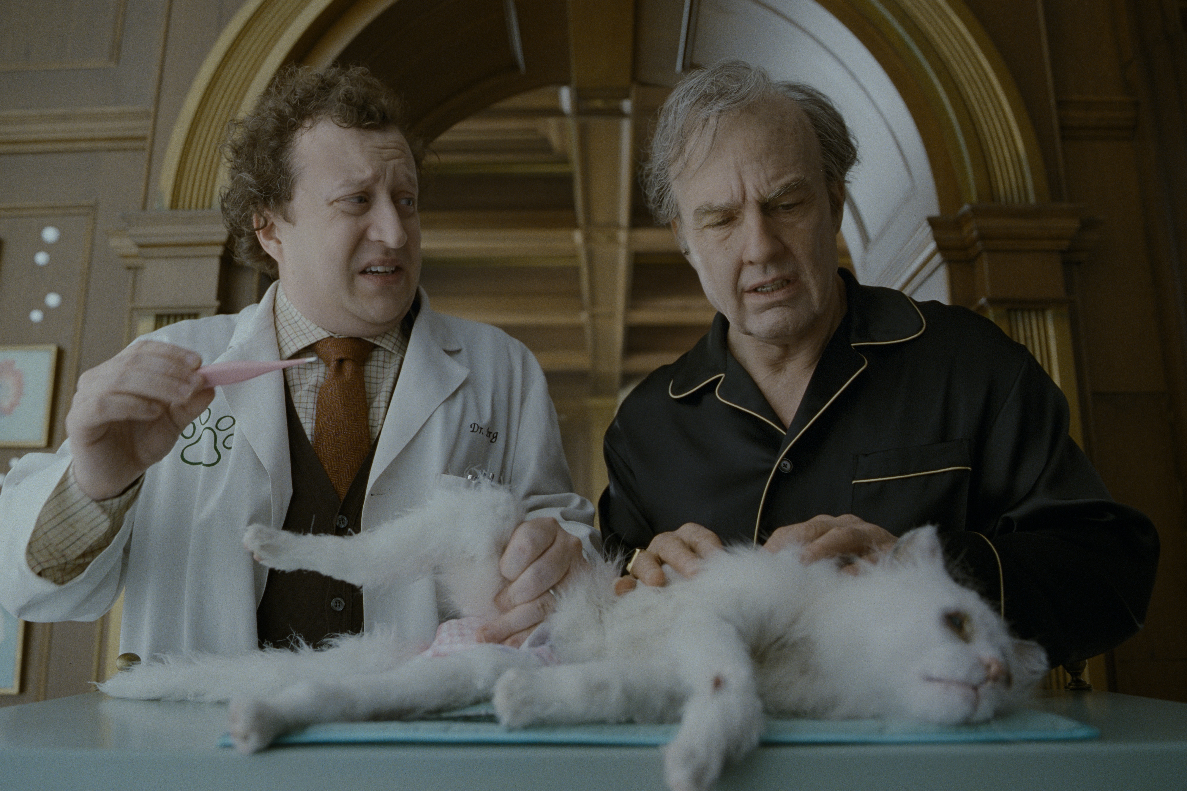 Two men, one wearing a vet's white lab coat and holding a thermometer, stand over an anamatronic white cat laid out on a table.