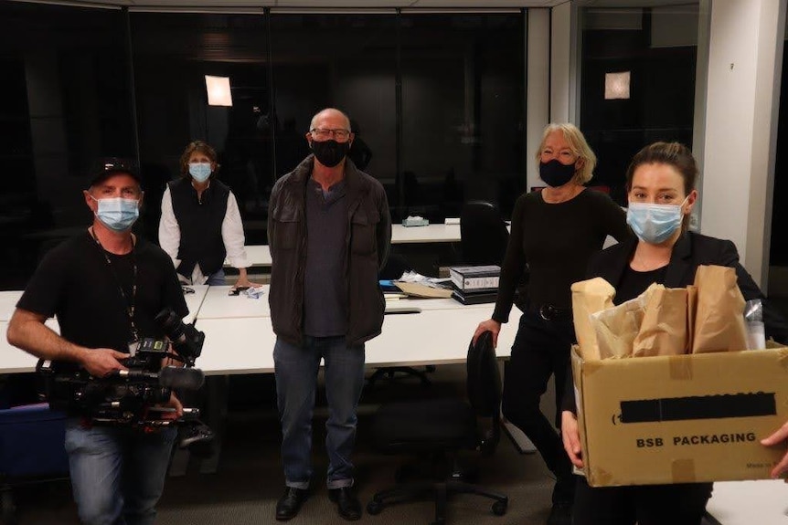 Five people wearing masks standing apart in office and police officer holding evidence box.