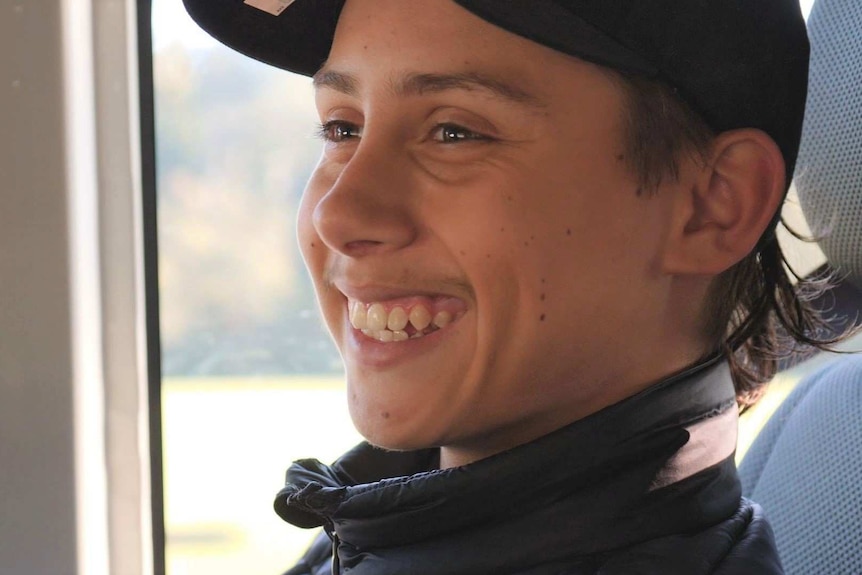 A teenage boy wearing a black cap grins widely as he sits on a bus.
