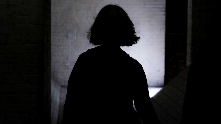 A young woman stands in profile in a darkened hallway near a room.