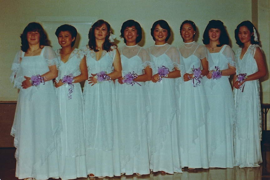 A group of women in white dresses stand in a row holding bouquets.