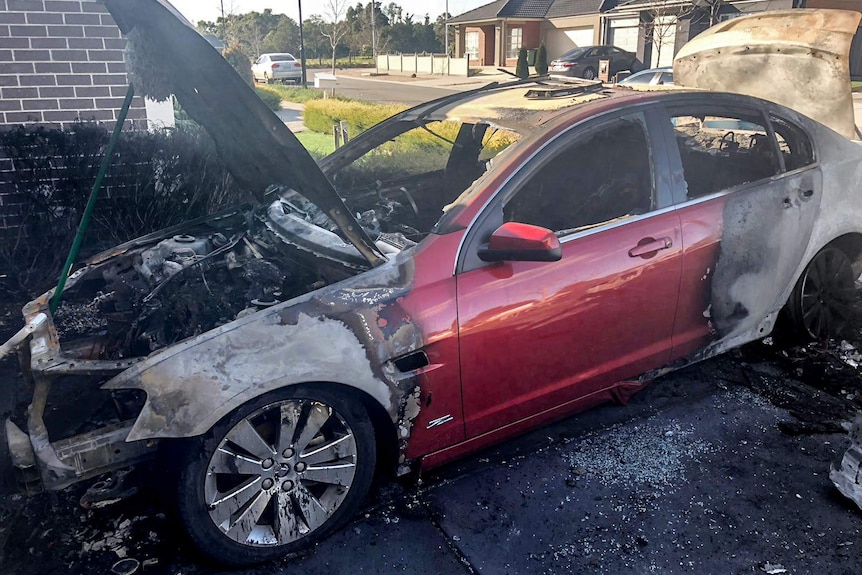 A burnt out Holden chassis sits in a driveway, with suburban homes behind.