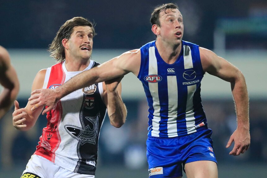 A male AFL player stands in front of another male AFL player, holding him back with his right arm.