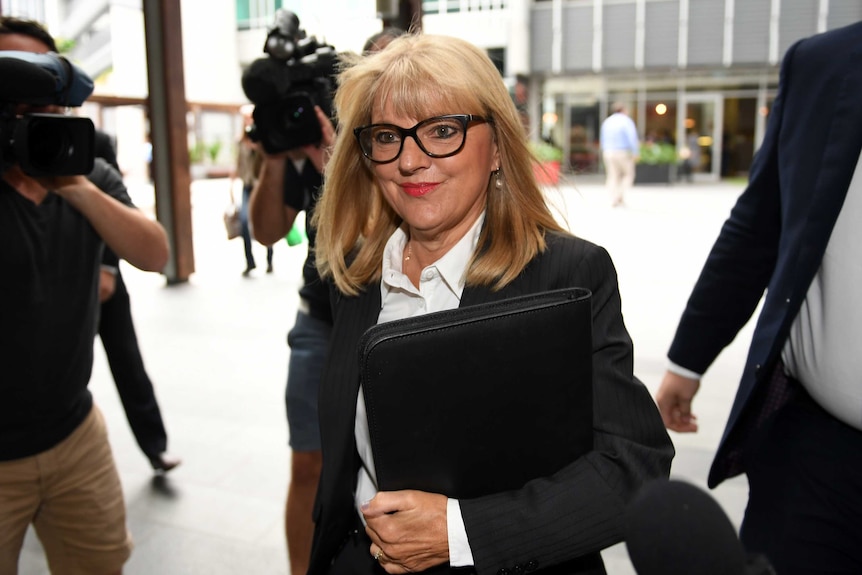 Donna Gates walks through a waiting media pack wearing a suit and carrying a brief case.