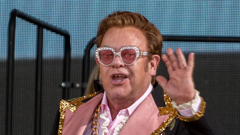 Sir Elton John standing on a concert stage with left hand close to his ear.