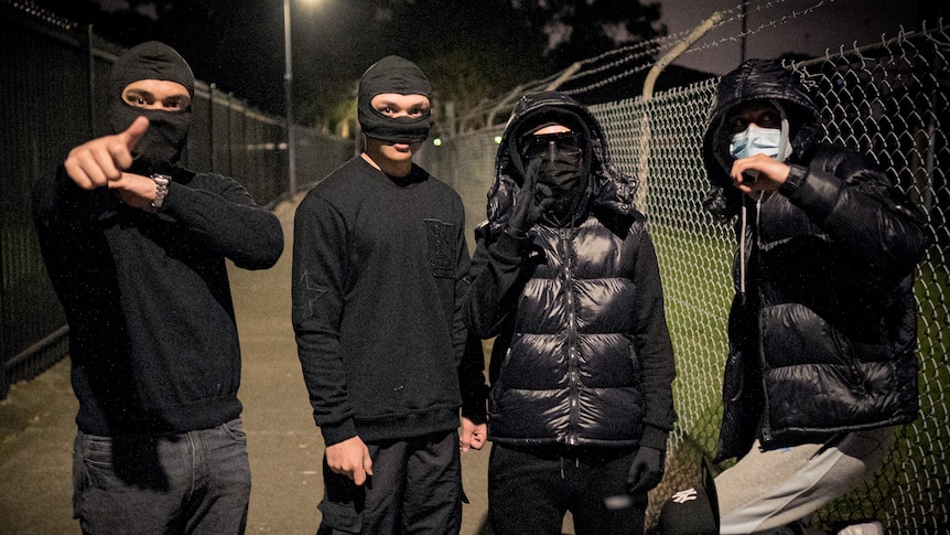 Four young people wearing black stand in a row, two wear balaclavas, the others wear masks and hoods. Three make hand signals.