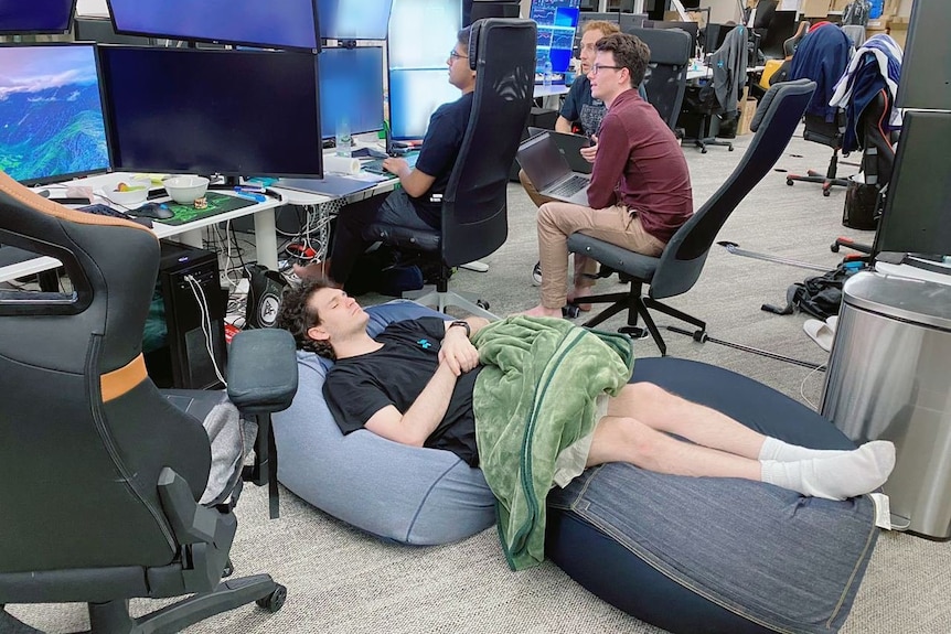 A young man sleeping on the floor of an office while other men work around him
