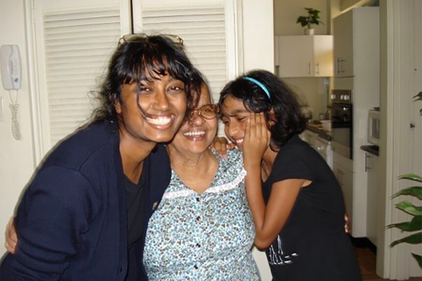 An adult Dinalie smiles as she hugs her grandmother and cousin
