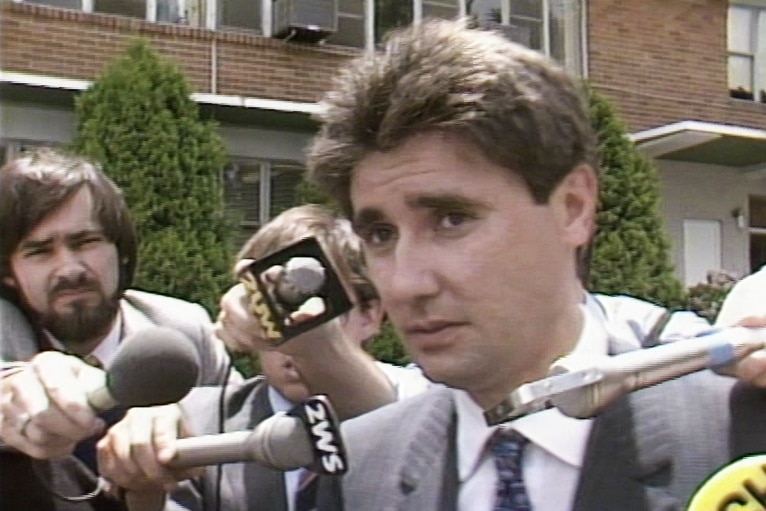 A man with brown hair and a grey suit surrounded by journalists holding microphones to his face.