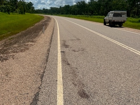 Dark blood stains on an empty road. There is a car parked on the side of the road. 