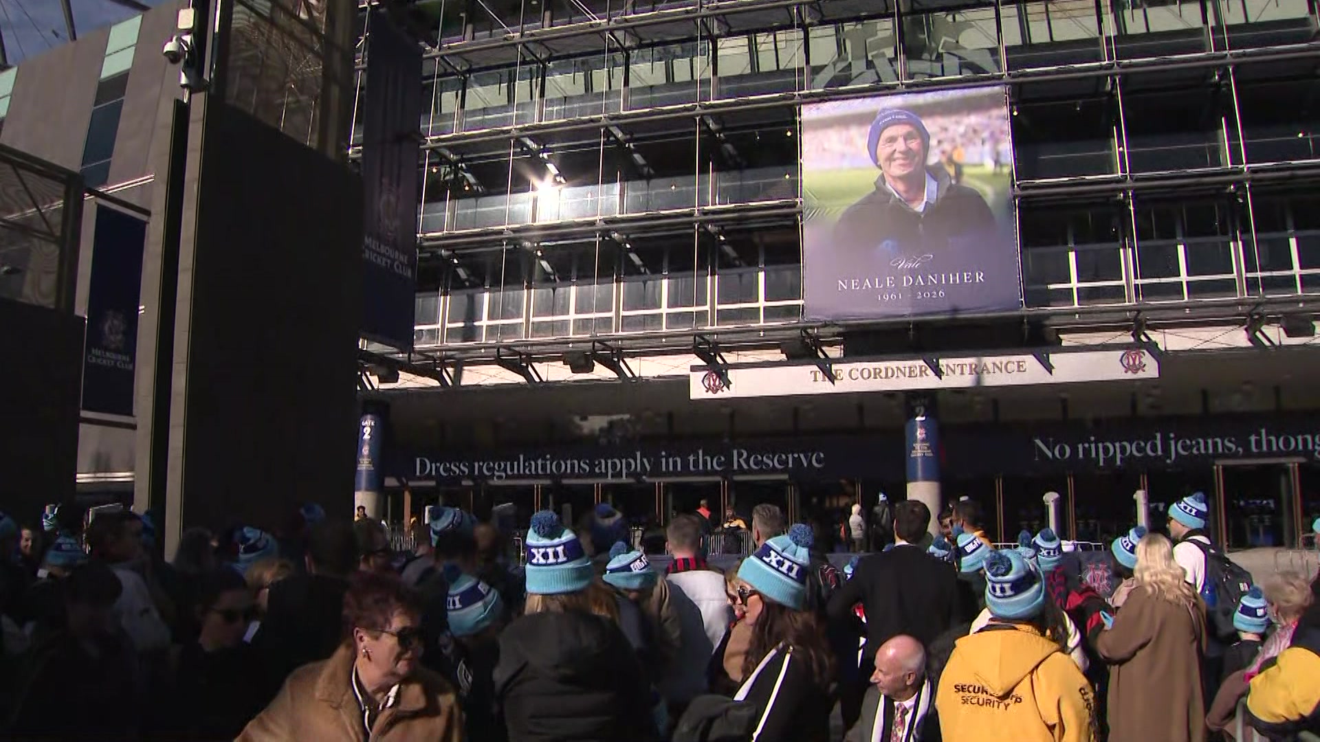 A crowd, many wearing light blue beanies, stands underneath a stadium billboard showing a smiling man wearing a beanie.