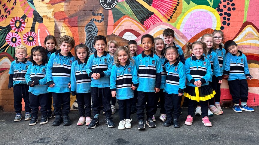 group of preschool children in blue uniforms pose in front of a very colourful mural