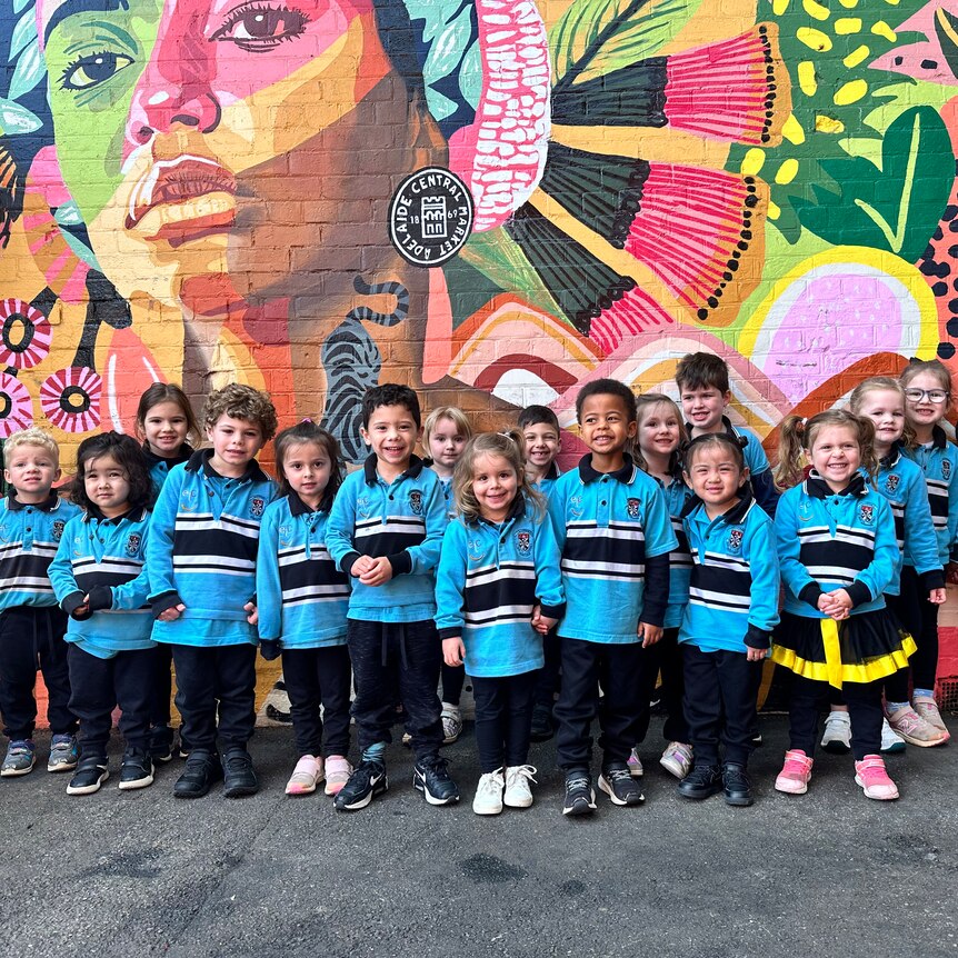 group of preschool children in blue uniforms pose in front of a very colourful mural