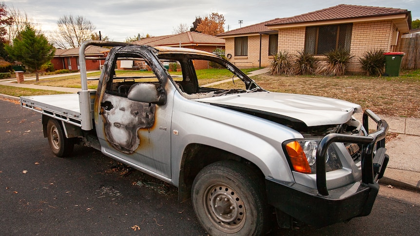 A burnt-out ute in front of a row of suburban houses.