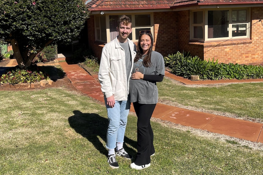 Roisin and Zac Tarrant outside their brick house in the sun. Roisin is pregnant and rests her hand on her bump