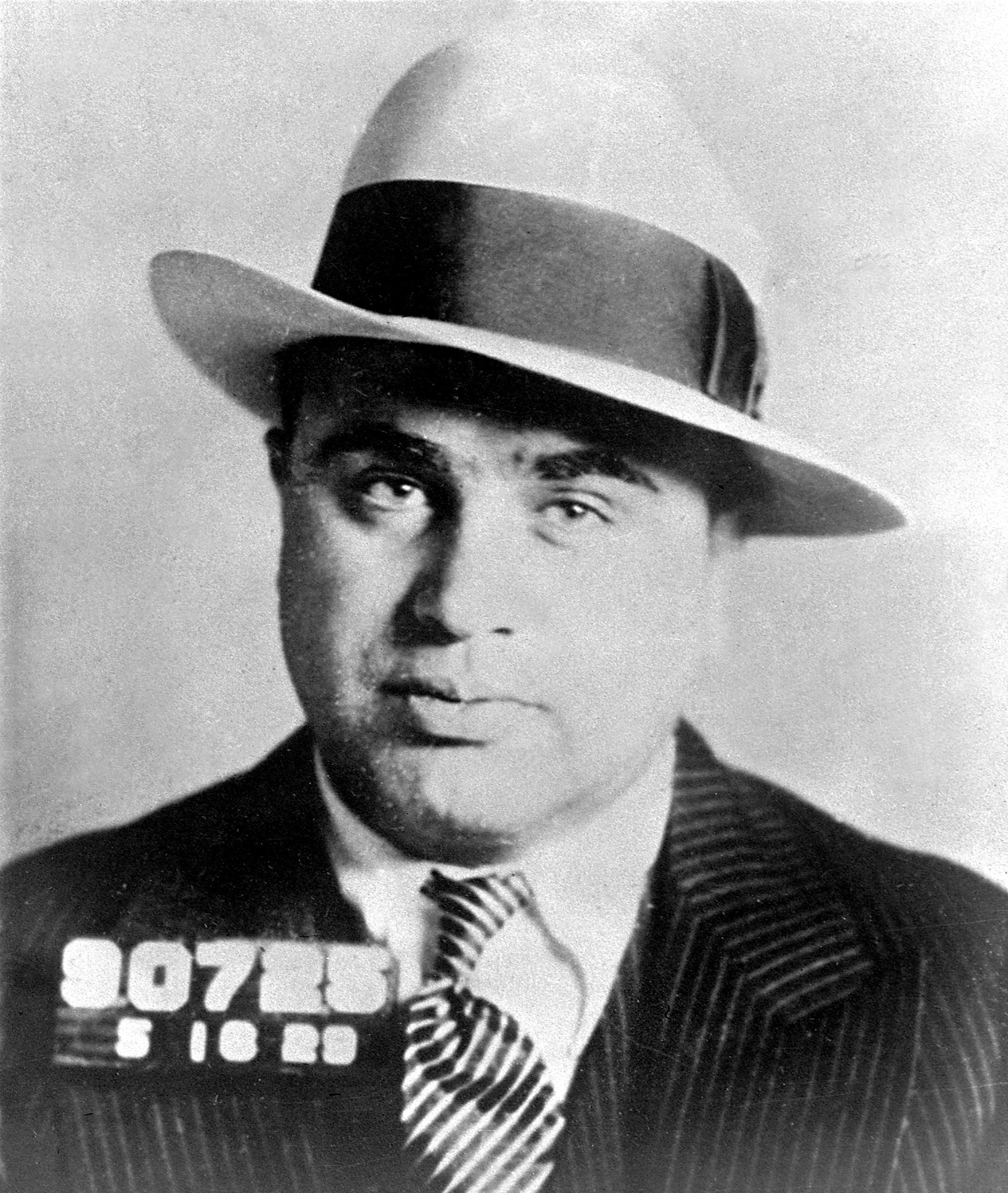 A black and white mugshot of a man wearing a fedora titled to the side and a slick suit.