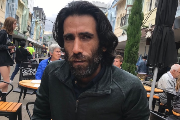 A man with long dark hair and beard sits at an outdoor cafe