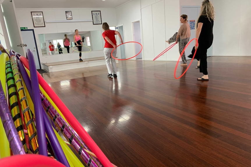 Hula hoop class with instructor on elevated platform three participants and stack of hula hoops against wall