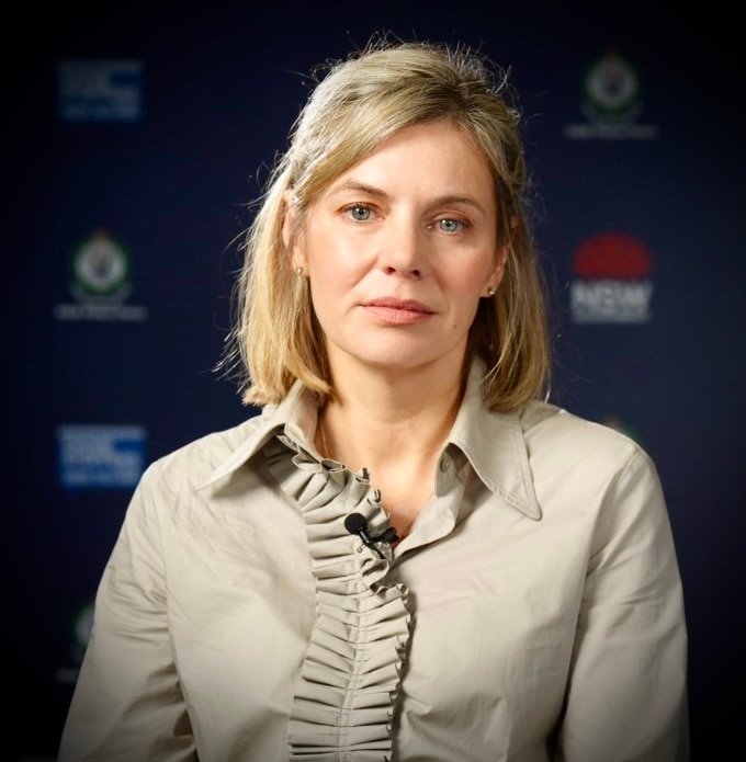 Kristen Hilton up close, with blonde hair, a beige top, with a serious expression, sitting in front of a NSW police media wall.