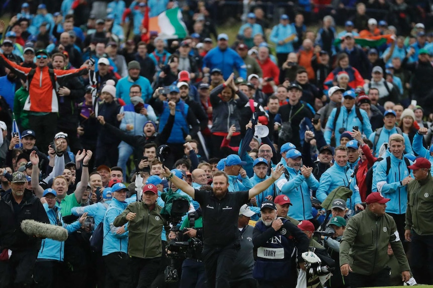 A large crowd celebrates, an Irish flag flies and Shane Lowry stands in front of it all with his arms raised.