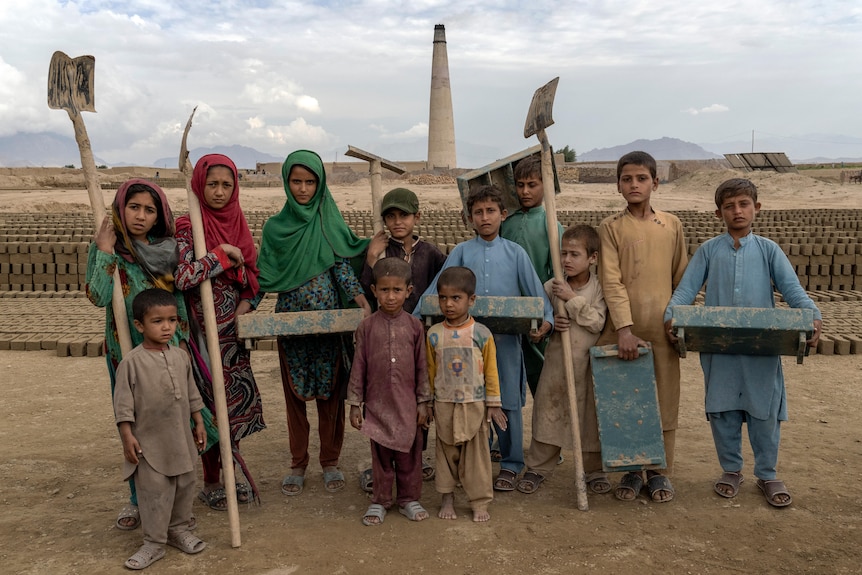 A group of Afghan children pose for a photo ata  brick factory, some holding shovels and other implements