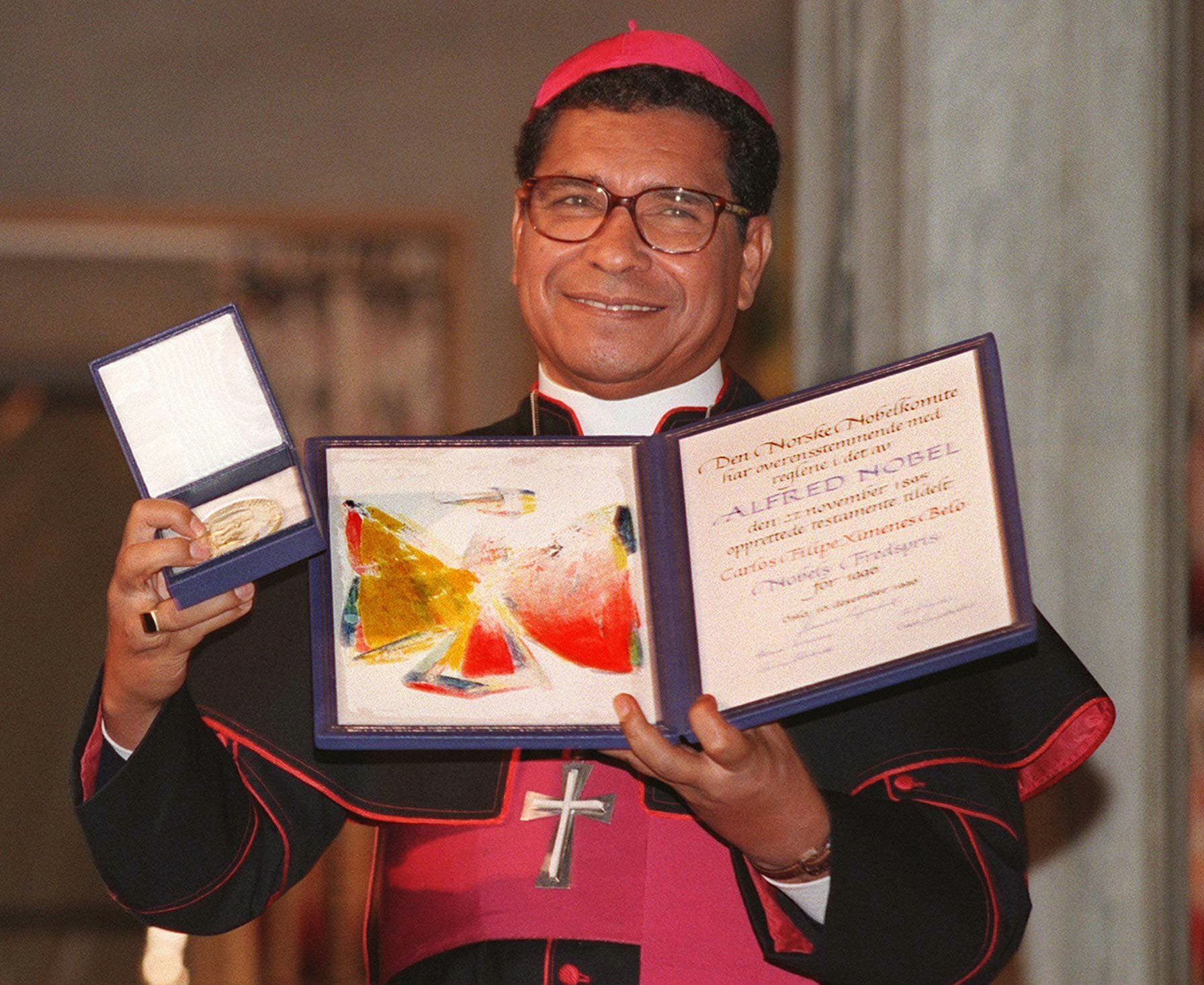 bishop smiling holding medal and certificate, wears red and black robes and glasses