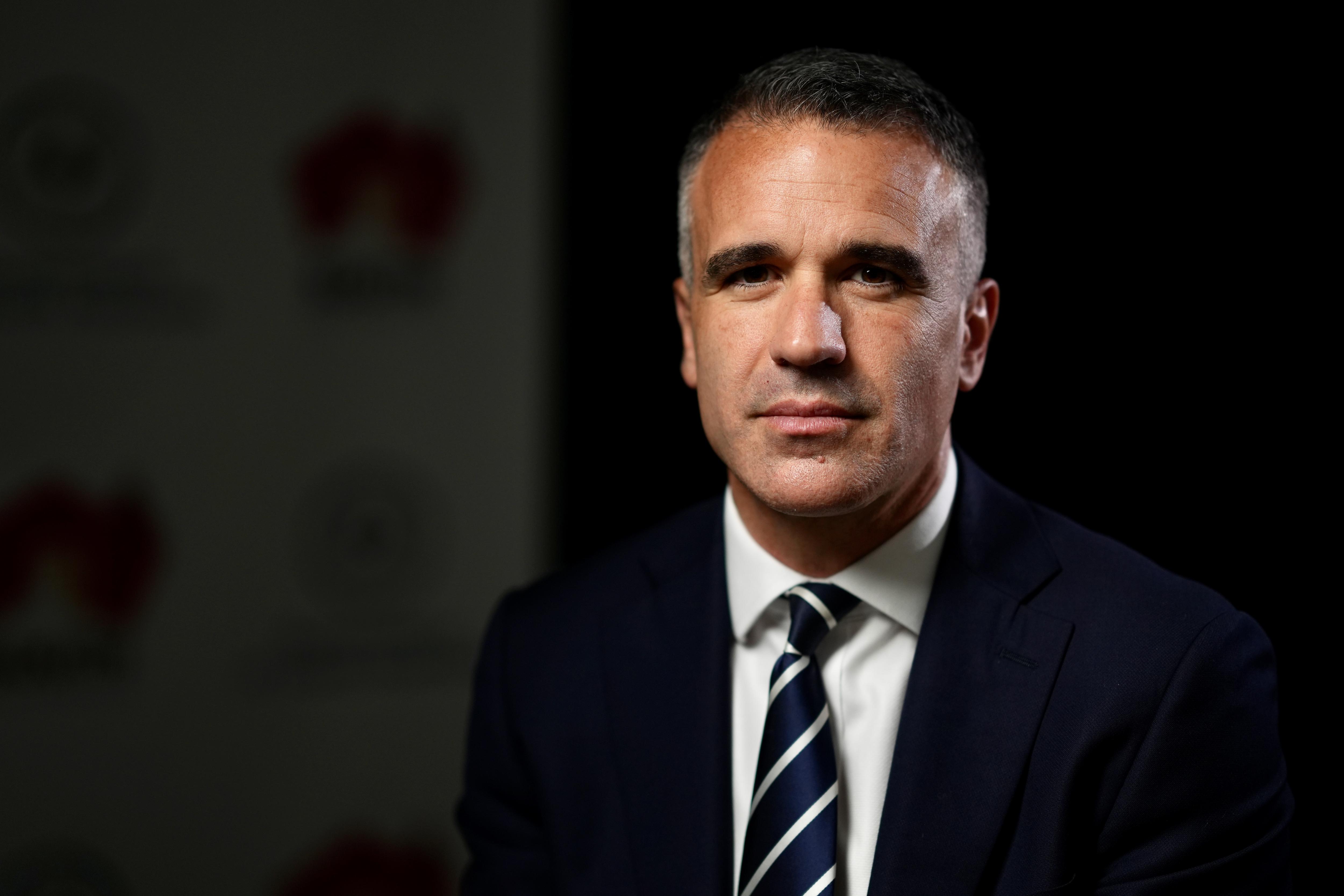 Close de um homem com uma expressão séria olhando diretamente para a câmera. Ele veste terno azul marinho, gravata e camisa branca.