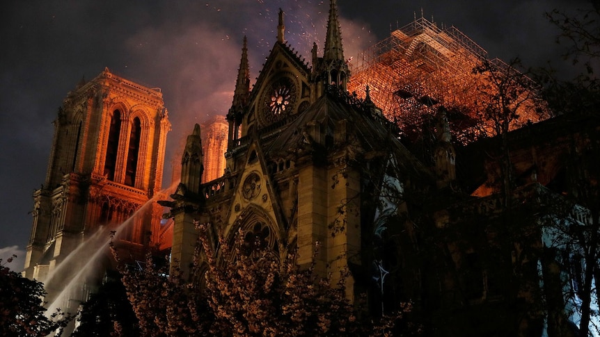Firefighters spray water onto a burning cathedral as embers drift across a dark sky