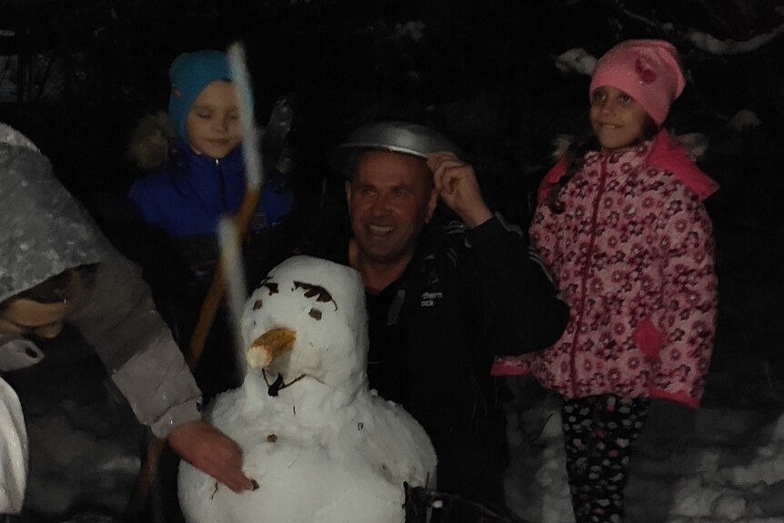 A man playing in the snow with a snowman and two children