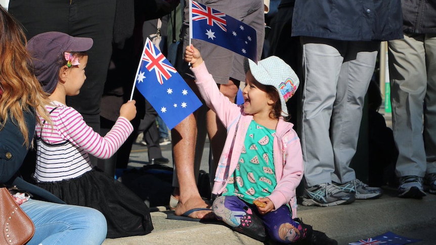 Two young girls holding small Australian flags sit on a footpath at the feet of adults.