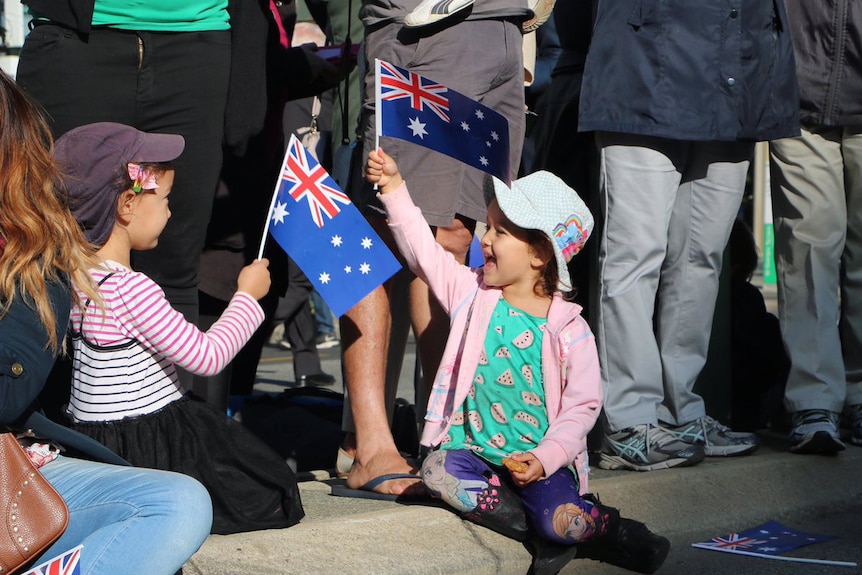 Two young girls holding small Australian flags sit on a footpath at the feet of adults.