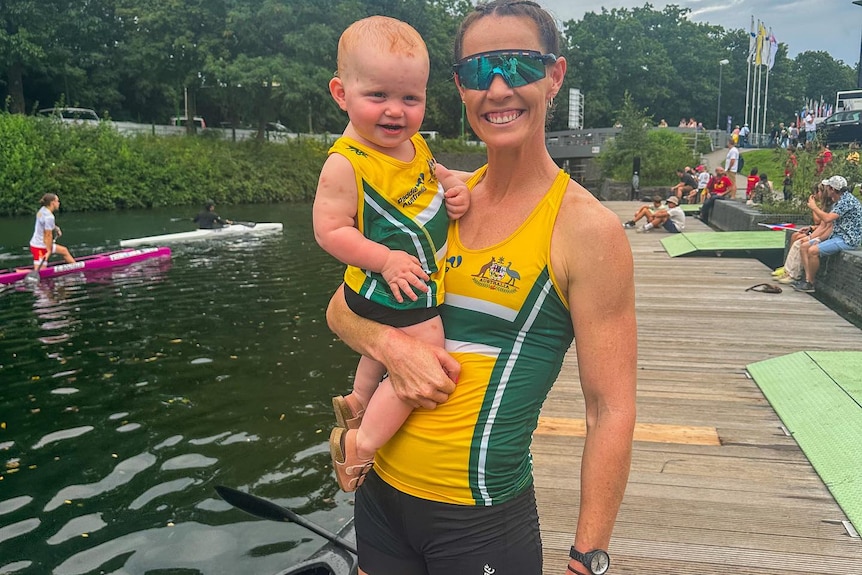 Alyce Wood wearing an Australian kayak uniform stands on a jetty and smiles while holding baby daughter Florence.
