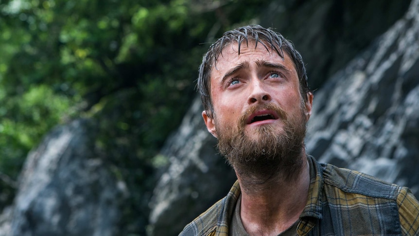 A bearded young man with wet hair stares upwards, rocks and foliage in background.