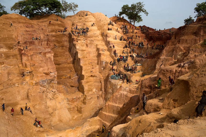 An open cut mine in Africa, with yellow rock hacked in steps, and people sitting all around