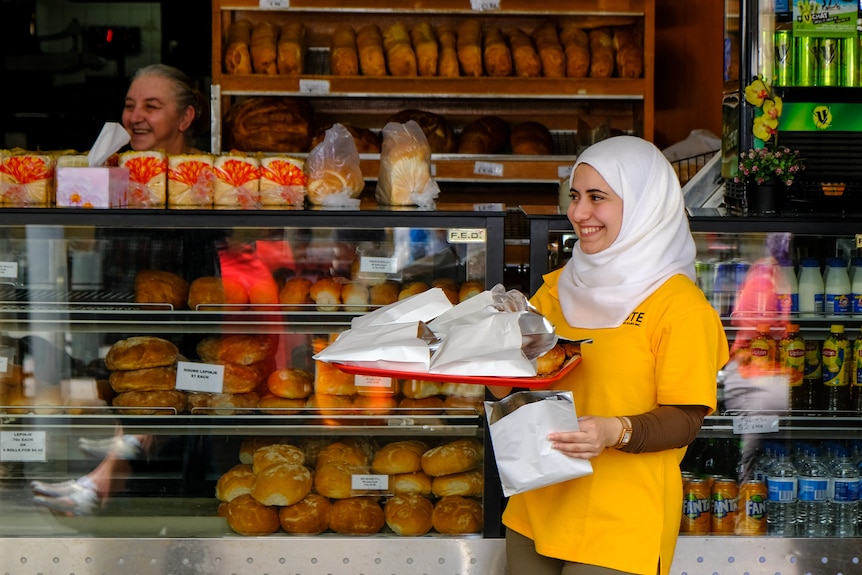 A woman with a yellow t-shirt wearing a white hair covering holds a tray of pastries in white bags